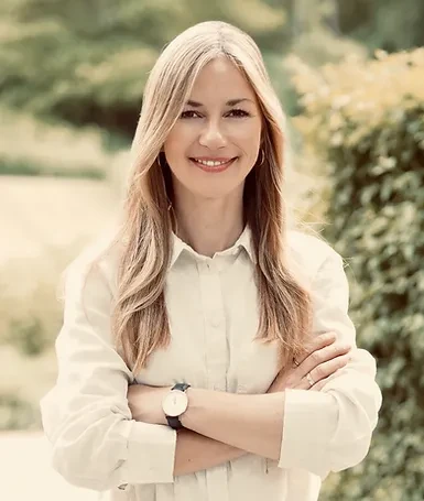 Junge Frau mit langen blonden Haaren lächelt selbstbewusst im Freien mit verschränkten Armen.